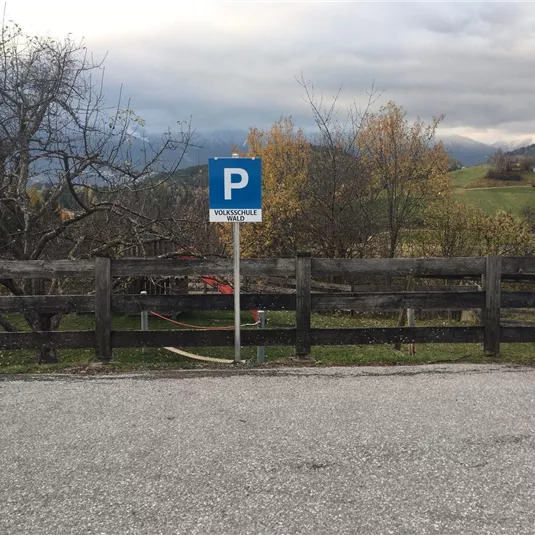 A parking sign stands by a road, surrounded by a wooden fence. In the background, there are trees and hilly landscapes.