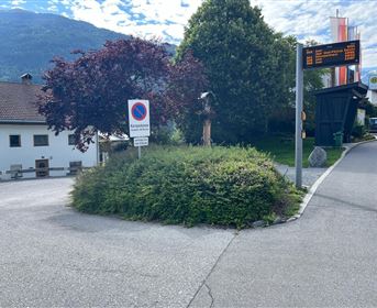 Ein Kreisverkehr mit einer grünen Bepflanzung und einem Baum. Im Hintergrund sind eine Straßenanzeige und einige Häuser sichtbar.