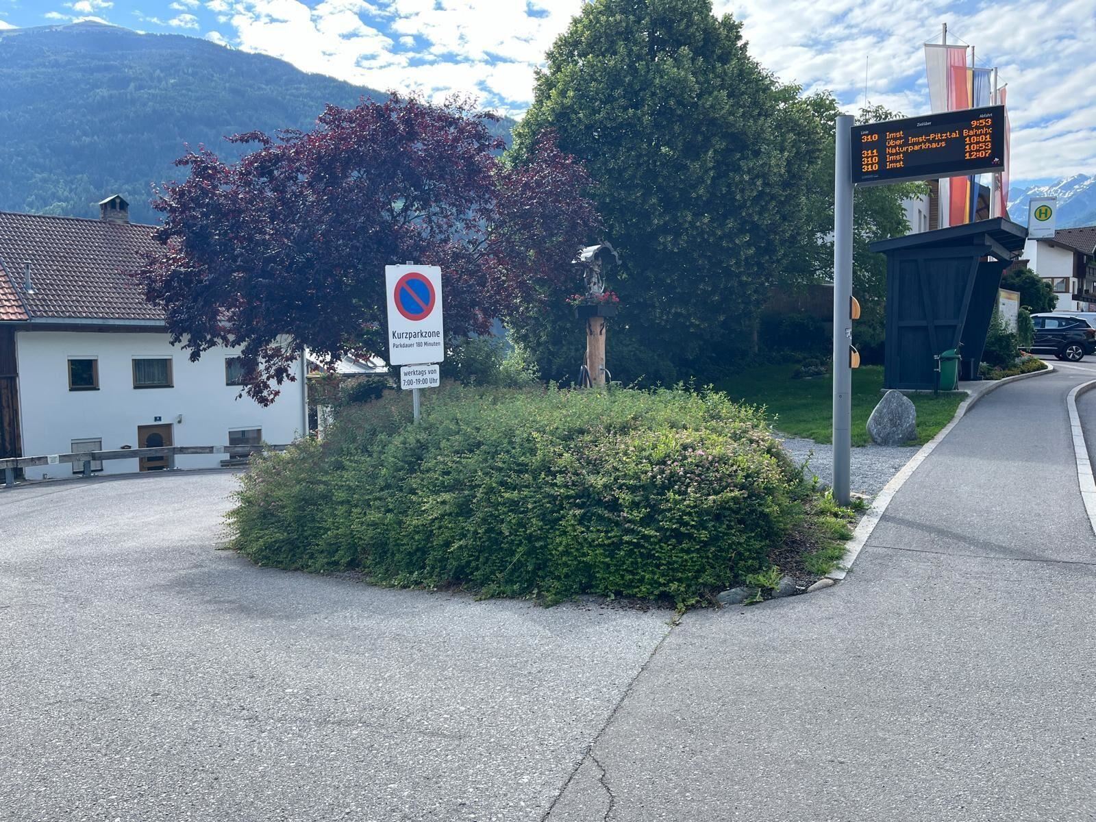 Ein Kreisverkehr mit einer grünen Bepflanzung und einem Baum. Im Hintergrund sind eine Straßenanzeige und einige Häuser sichtbar.