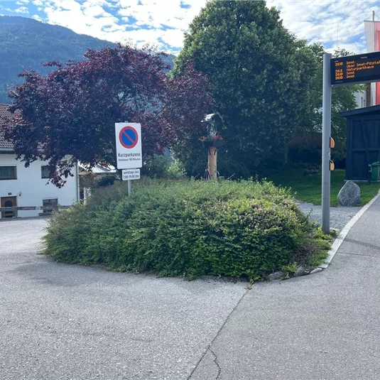 Ein Kreisverkehr mit einer grünen Bepflanzung und einem Baum. Im Hintergrund sind eine Straßenanzeige und einige Häuser sichtbar.