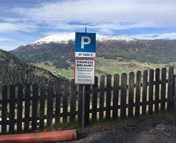 A parking sign in the mountains with a beautiful view of snow-capped peaks and green valleys. The area is surrounded by a wooden fence.