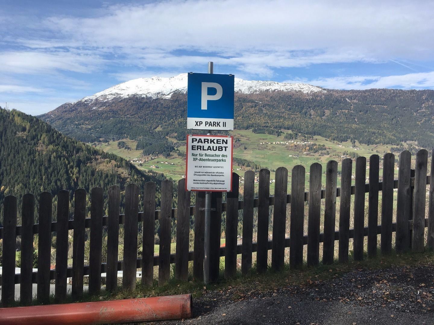 A parking sign in the mountains with a beautiful view of snow-capped peaks and green valleys. The area is surrounded by a wooden fence.
