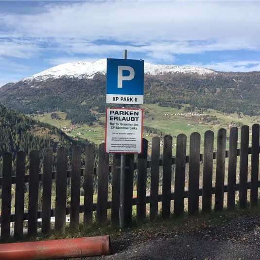 A parking sign in the mountains with a beautiful view of snow-capped peaks and green valleys. The area is surrounded by a wooden fence.