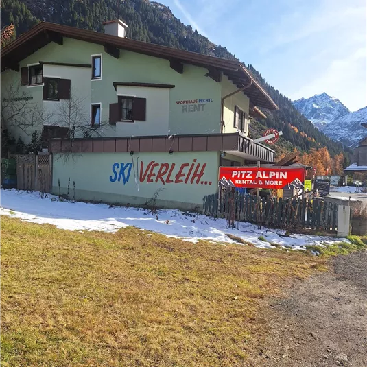 Ein Gebäude in den Bergen mit einem großen Schild für einen Skiverleih. Im Vordergrund liegt eine schneebedeckte Wiese.