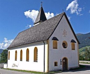 Eine kleine Kirche mit einem Spitzdach und weißen Wänden. Sie liegt in einer ländlichen Umgebung unter einem blauen Himmel.