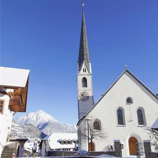 Eine malerische Kirche mit einem hohen Turm im Winter. Der Hintergrund zeigt schneebedeckte Berge unter einem klaren blauen Himmel.