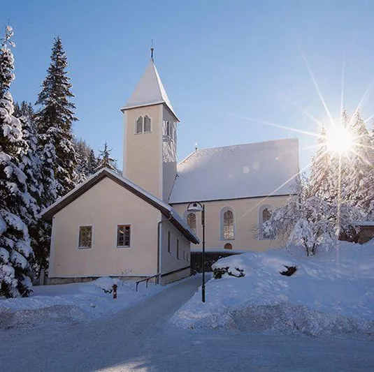Eine idyllische Kirche umgeben von schneebedeckten Bäumen. Die Sonne strahlt hell hinter der Kirche und sorgt für eine warme Atmosphäre.