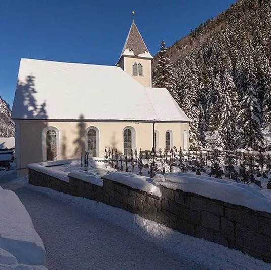 Eine kleine Kirche im Schnee, umgeben von hohen Tannenbäumen. Der Himmel ist klar und blau, was eine ruhige Winterlandschaft schafft.