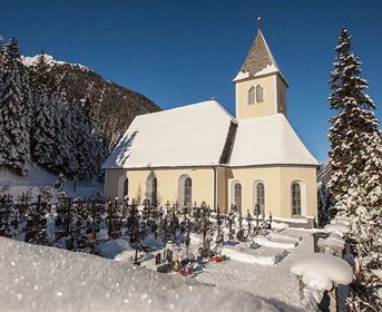 Eine kleine Kirche im Schnee, umgeben von hohen Tannen. Der Himmel ist klar und blau, was eine ruhige Atmosphäre schafft.