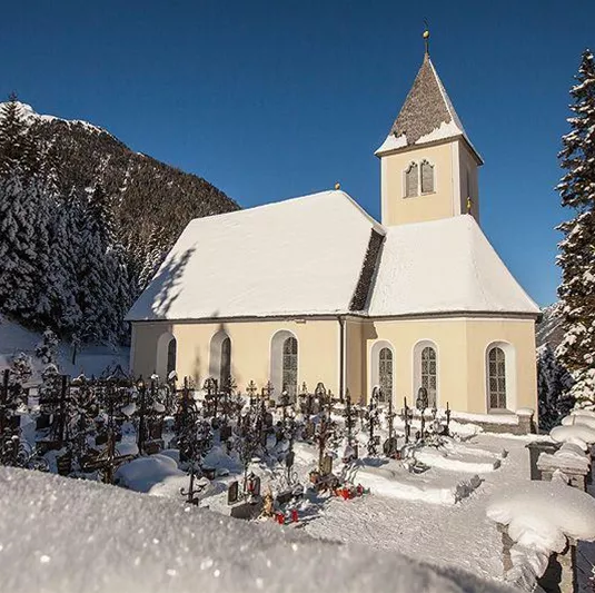 Eine kleine Kirche steht in einer verschneiten Landschaft. Umgeben von Tannenbäumen und einem Friedhof ist die Szene ruhig und malerisch.