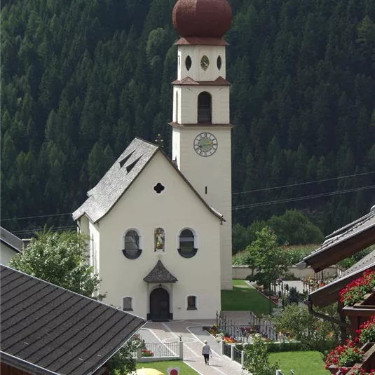 Eine malerische Kirche mit einem charakteristischen Zwiebelturm in einer grünen Landschaft. Menschen versammeln sich vor der Kirche.