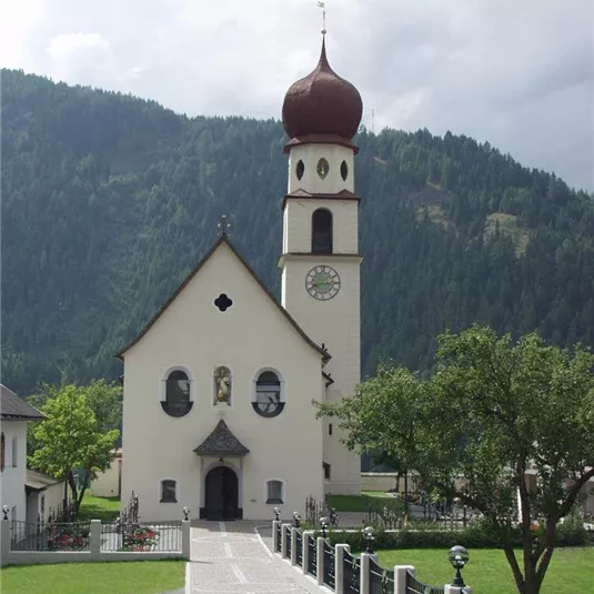 Eine malerische Kirche mit einem hohen Glockenturm und einer charakteristischen Zwiebelturmspitze. Im Hintergrund sind grüne Hügel und ein bewölkter Himmel zu sehen.