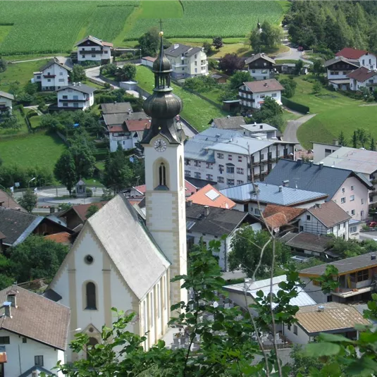 Ein Blick auf eine malerische, ländliche Stadt mit zahlreichen Häusern und einer auffälligen Kirche im Vordergrund. Die Umgebung ist grün und von Hügeln geprägt.