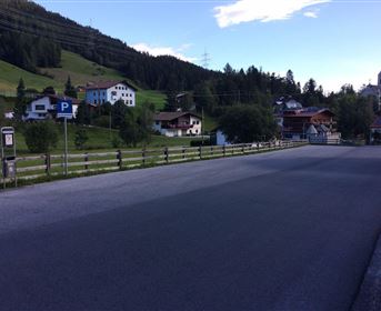 Eine ruhige Straße in einem Dorf mit traditionellen Häusern im Hintergrund. Die umliegenden Berge sind grün und es ist ein klarer Himmel zu sehen.