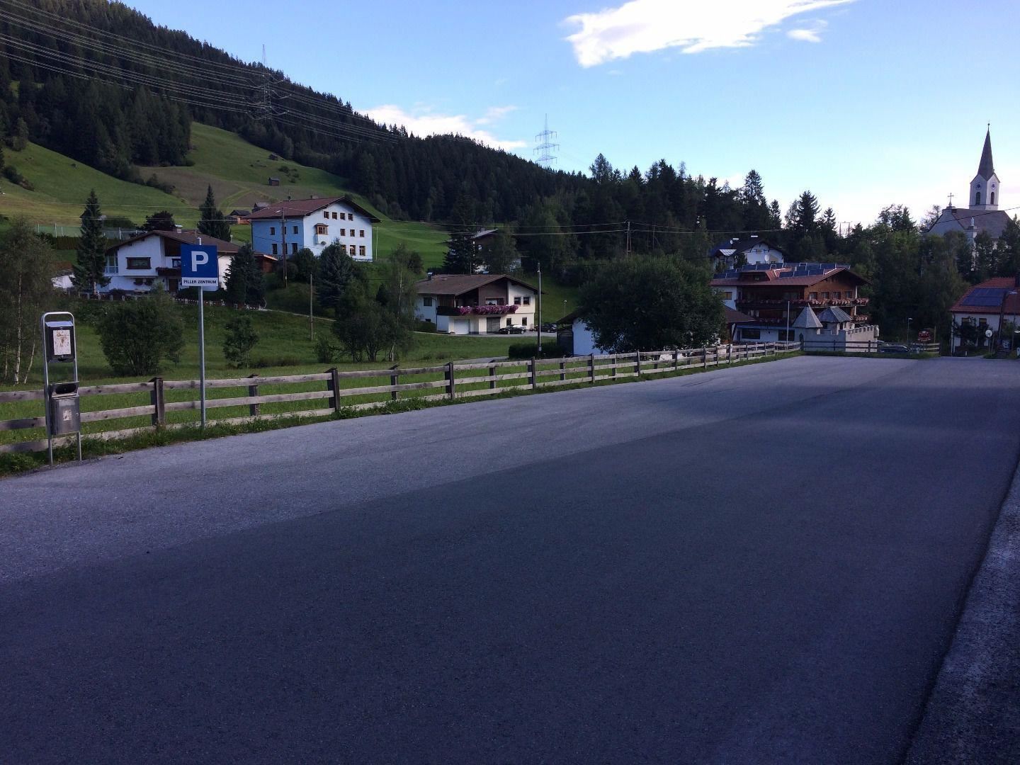 A quiet street in a village with traditional houses in the background. The surrounding mountains are green and a clear sky can be seen.