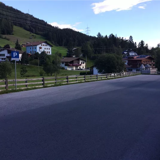 A quiet street in a village with traditional houses in the background. The surrounding mountains are green and a clear sky can be seen.