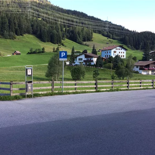 A peaceful landscape with green meadows and a parking lot. In the background, several traditional houses and trees can be seen.