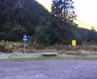 Ein Parkplatz mit einem blauem P-Schild und einer Holzbank. Im Hintergrund sind Bäume und Berge zu sehen.