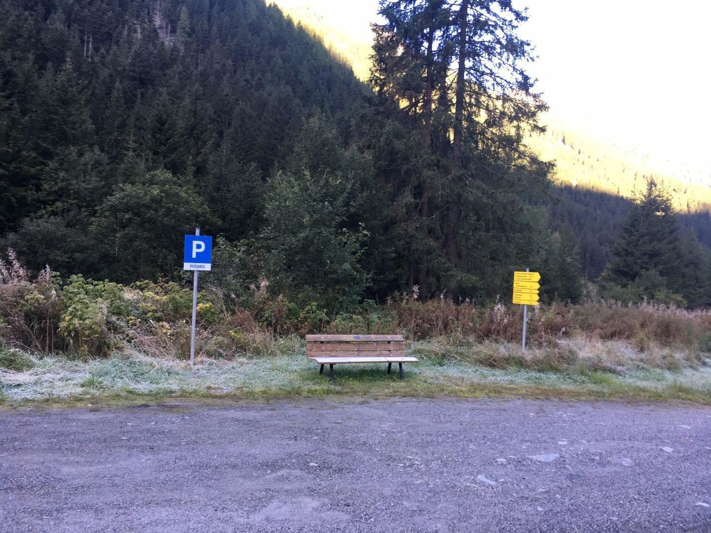 Ein Parkplatz mit einem blauem P-Schild und einer Holzbank. Im Hintergrund sind Bäume und Berge zu sehen.