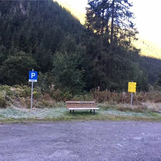 Ein Parkplatz mit einem blauem P-Schild und einer Holzbank. Im Hintergrund sind Bäume und Berge zu sehen.