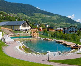 A beautiful natural pool surrounded by green meadows and mountains. In the background, you can see a cozy building and a few sun loungers.