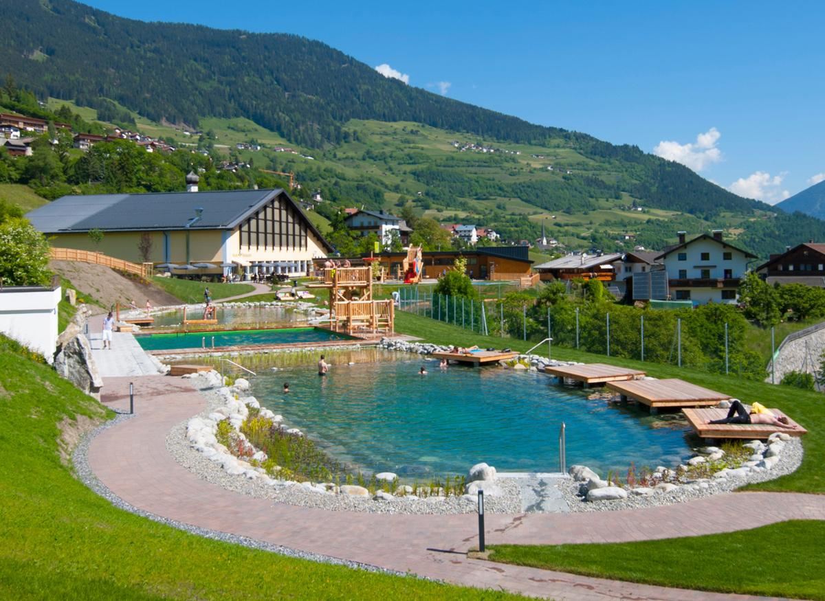 Ein schöner Naturpool umgeben von grünen Wiesen und Bergen. Im Hintergrund sieht man ein gemütliches Gebäude und einige Sonnenliegen.