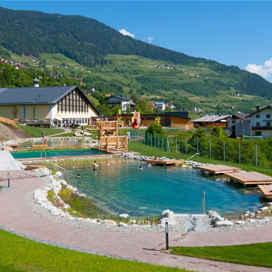 Ein schöner Naturpool umgeben von grünen Wiesen und Bergen. Im Hintergrund sieht man ein gemütliches Gebäude und einige Sonnenliegen.