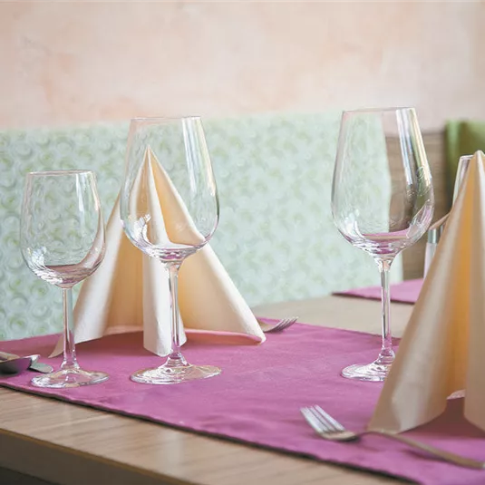 An elegantly set table with wine glasses and napkins shaped like lace. The tablecloth is in purple and the background is in soft shades of green.