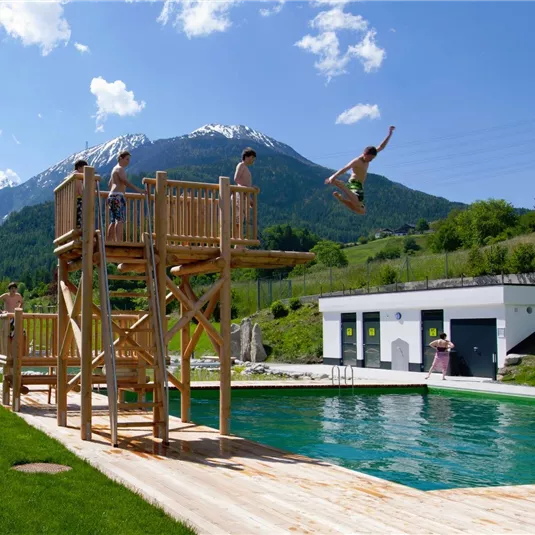 Ein schöner Schwimmplatz mit einem Holzspringturm und einer grünen Wiese. Im Hintergrund sind Berge mit schneebedeckten Gipfeln und ein blauer Himmel zu sehen.