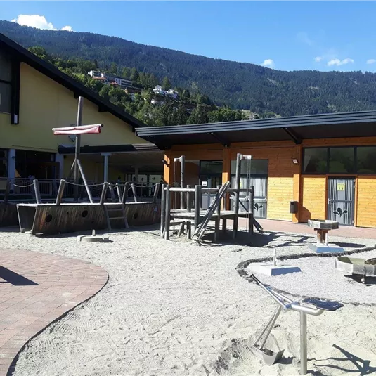 Ein Spielplatz mit Sand, Rutschen und Spielgeräten. Im Hintergrund sind Gebäude und Berge sichtbar.
