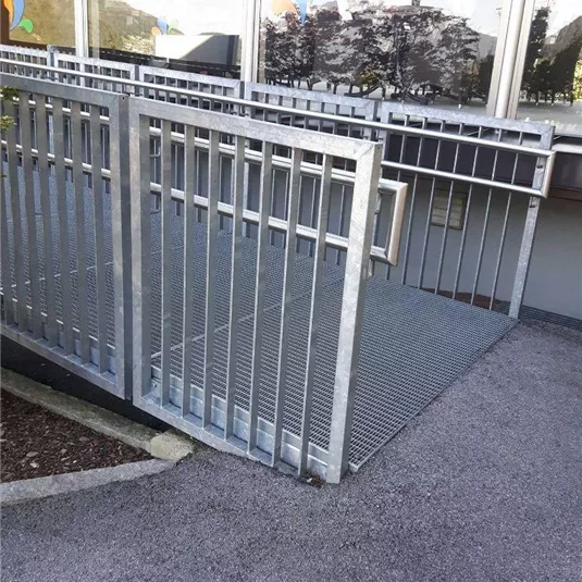 Eine Rampe aus Metall mit einem rutschfesten Gitterboden. Sie führt zu einem Gebäude mit großen Fenstern.