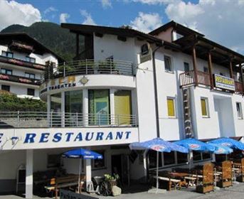 Ein modernes Restaurantgebäude mit einer einladenden Terrasse. Im Hintergrund sind mehrere Wohngebäude und ein klarer Himmel zu sehen.