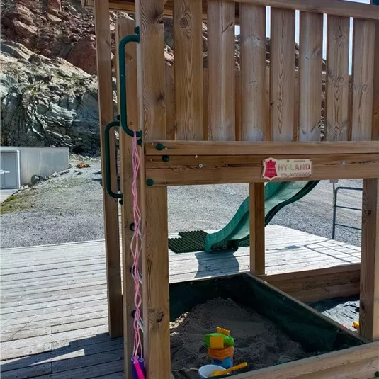 Ein Holzspielplatz mit einer Sandkiste und einer Rutsche. Im Sand befinden sich einige Spielzeuge, während im Hintergrund Felsen sichtbar sind.