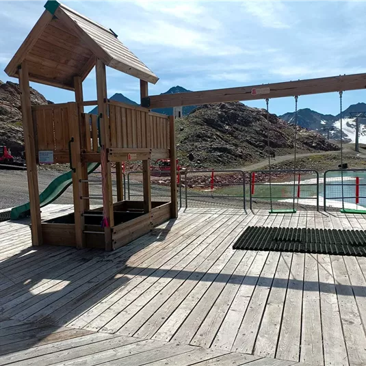 Ein Spielplatz mit einer Holzhütte und einer Schaukel steht vor einer bergigen Landschaft. Im Hintergrund sind schneebedeckte Berge und ein blauer Himmel zu sehen.