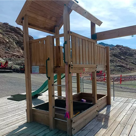 Ein Spielplatz aus Holz mit einer Rutsche und Sandkasten. Im Hintergrund sind felsige Hügel und ein blauer Himmel zu sehen.