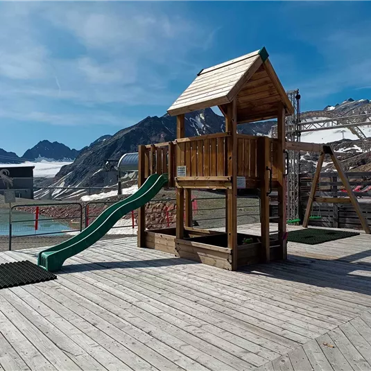 Ein Holzspielplatz mit einer Rutsche steht auf einem Holzdeck. Im Hintergrund sind Berge und ein blauer Himmel zu sehen.