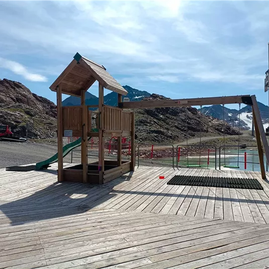 Ein Spielplatz aus Holz mit einer Rutsche und einem kleinen Haus steht auf einer Holzterrasse. Im Hintergrund sind Berge und ein See zu sehen.