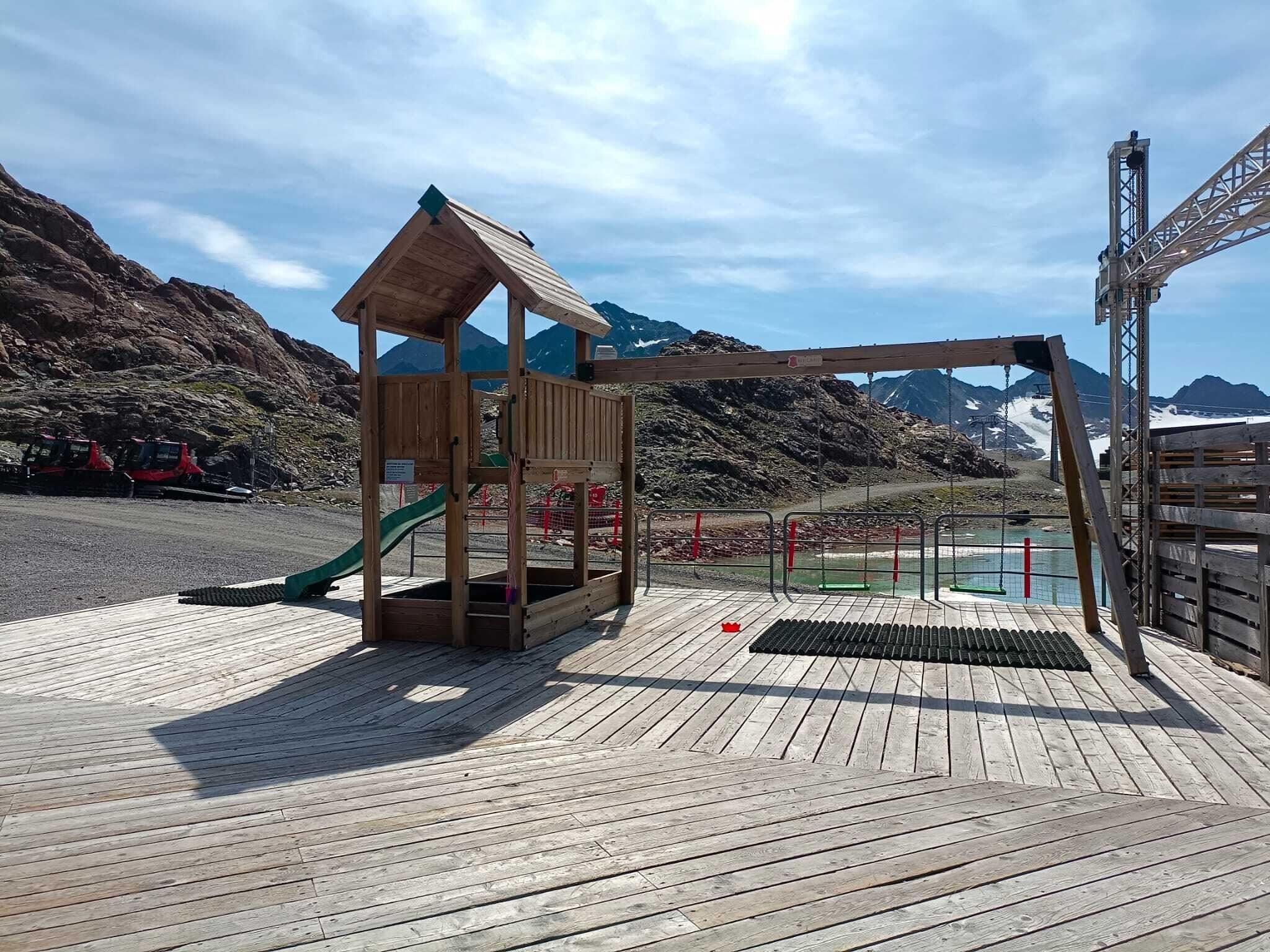 Ein Spielplatz aus Holz mit einer Rutsche und einem kleinen Haus steht auf einer Holzterrasse. Im Hintergrund sind Berge und ein See zu sehen.