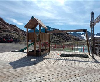 A playground made of wood with a slide and a small house stands on a wooden terrace. In the background, mountains and a lake can be seen.
