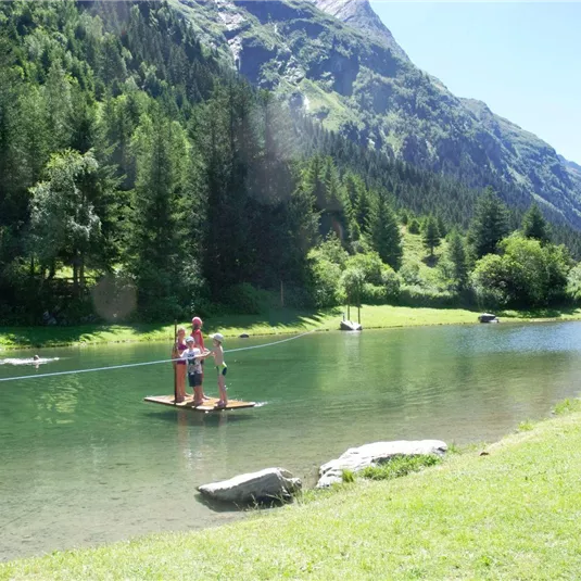 Two people are standing on a raft in a clear lake, surrounded by green forests and mountains. The sun is shining on a beautiful, clear day.