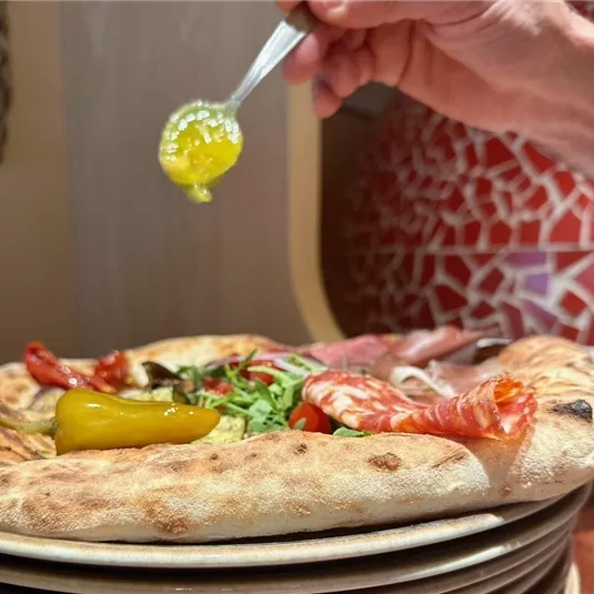 A fresh pizza with various toppings like salami and pepperoni. A green sauce is currently being added on top.