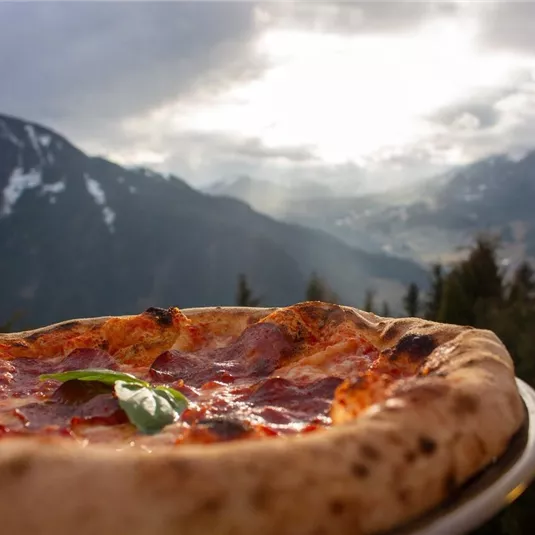 A delicious pizza with pepperoni and fresh basil in front of an impressive mountain landscape. The sky is cloudy, but the light creates a picturesque atmosphere.