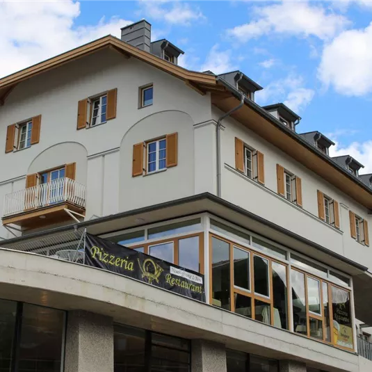 Ein modernes Gebäude mit einem hellen Fassadenanstrich und orangefarbenen Fensterläden. Im Erdgeschoss befindet sich ein Restaurant mit großen Fenstern.