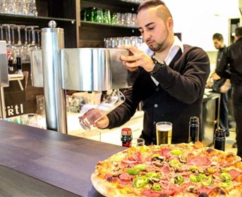 A waiter is tapping a beer at the bar, while a large pizza with various toppings is in the foreground. The atmosphere is lively and inviting.