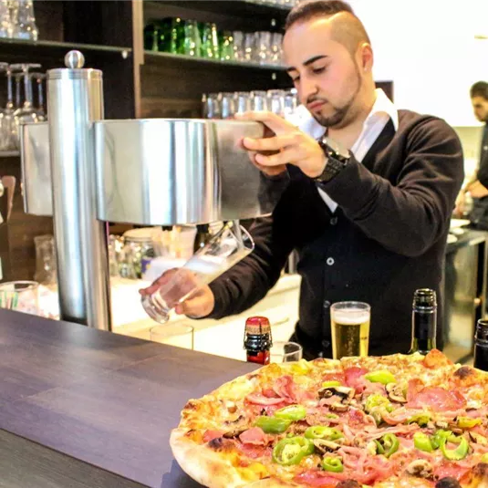 Ein Kellner zapft ein Getränk an der Bar, während eine große Pizza mit verschiedenen Belägen vor ihm steht. Im Hintergrund sind weitere Mitarbeiter und Gläser sichtbar.