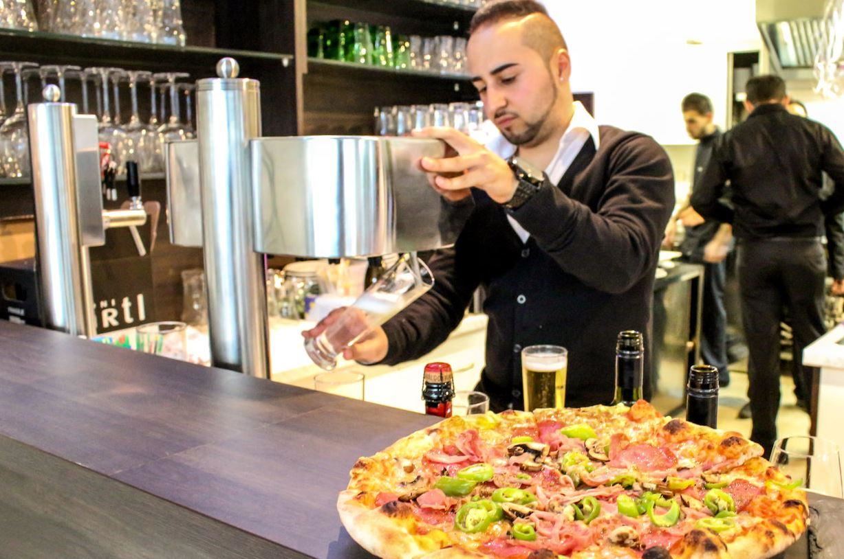 Ein Kellner zapft ein Getränk an der Bar, während eine große Pizza mit verschiedenen Belägen vor ihm steht. Im Hintergrund sind weitere Mitarbeiter und Gläser sichtbar.