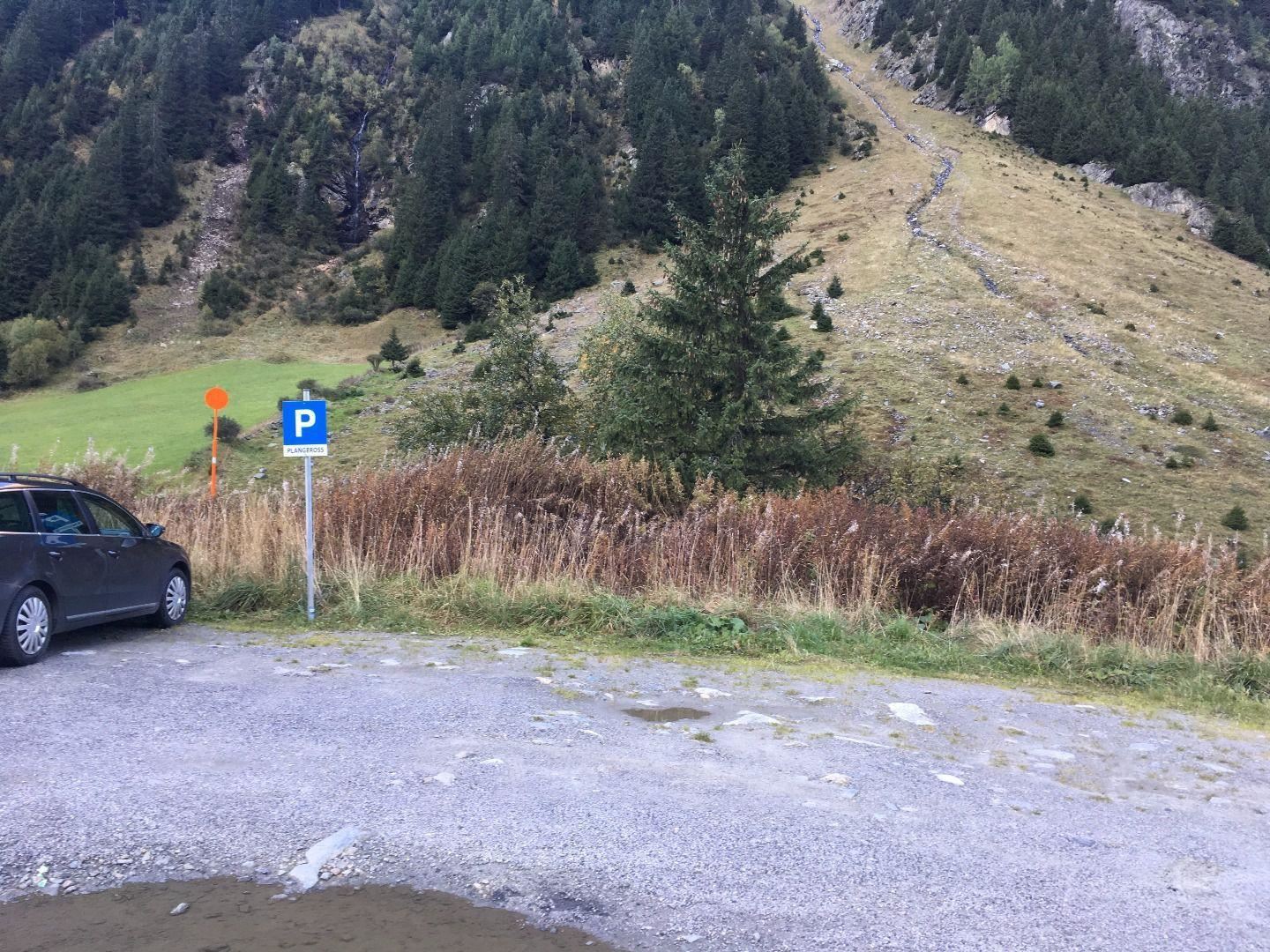 Ein Parkplatz am Fuße eines Hügels in der Natur. Im Hintergrund sind grüne Wiesen und hohe Bäume zu sehen.