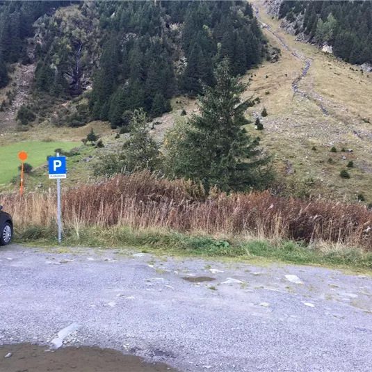Ein Parkplatz am Fuße eines Hügels in der Natur. Im Hintergrund sind grüne Wiesen und hohe Bäume zu sehen.