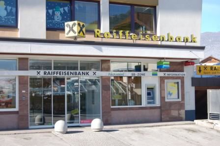 Die Raiffeisenbank mit großen Fenstern und einem klaren Entrée. Es gibt eine digitale Anzeigetafel und einen SB-Bereich vor dem Gebäude.