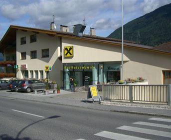 Ein modernes Bankgebäude mit Glasfront und einem großen Raiffeisenbank-Schild. Im Vordergrund sind mehrere geparkte Autos und ein Fußgängerüberweg zu sehen.